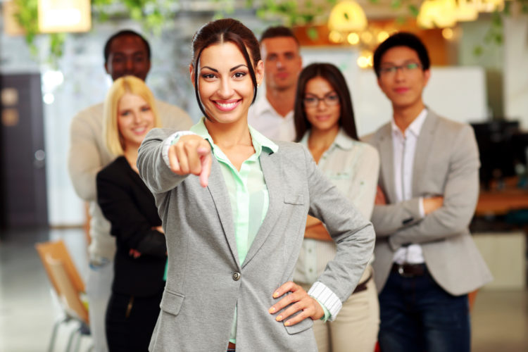 Smiling handsome businesswoman with team in office pointing finger at you
